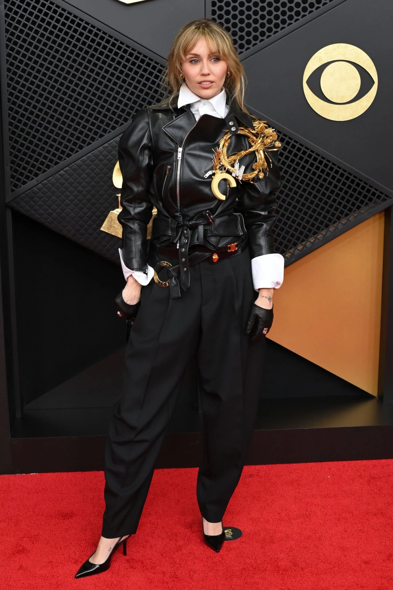Miley Cyrus poses on the Grammy red carpet in leather jacket, balloon trousers, and oversized gold MC brooch